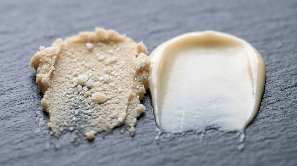 Macro close-up of two smears of shea butter on grey slate: the left one grainy and sandy with visible hard crystals, the right one perfectly smooth and glossy